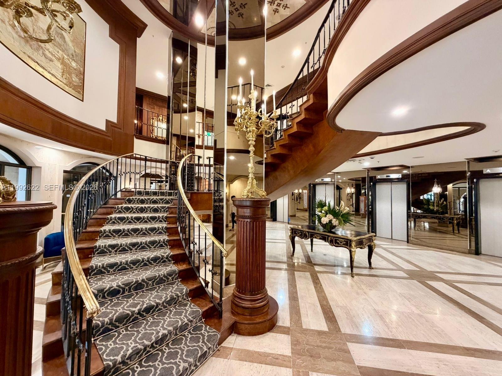A grand foyer features a curved staircase with patterned carpet and a central candelabra pedestal — 6039 Collins Ave # 1506, Miami Beach FL 33140, MIAMI BEACH, FL