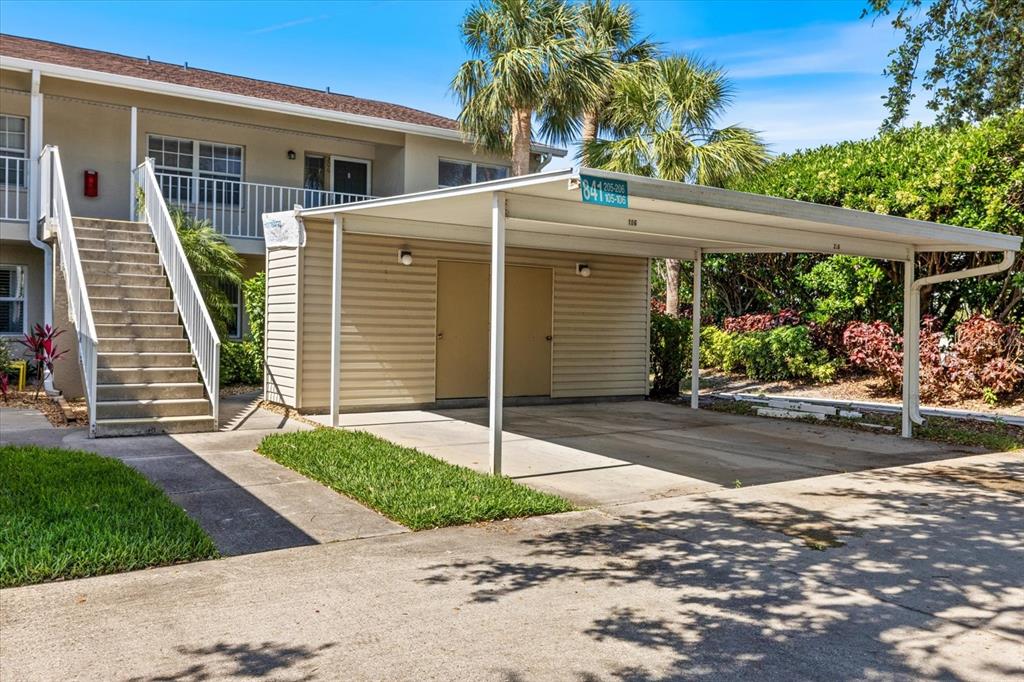An outdoor view of a covered carport with storage units next to an apartment building staircase — 841 WATERSIDE DRIVE UNIT 206, VENICE, FL