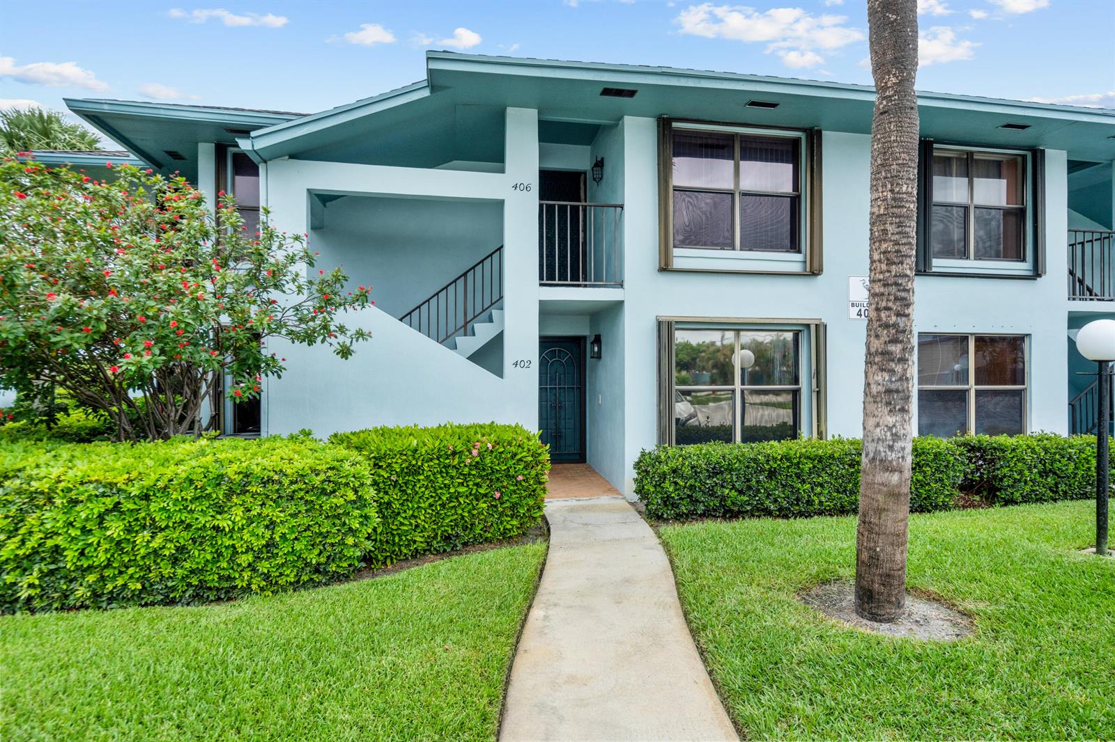 A two-story light blue apartment building features a concrete walkway, green hedges, and palm trees — 950 S Kanner Highway # 402, Stuart FL 34994, STUART, FL