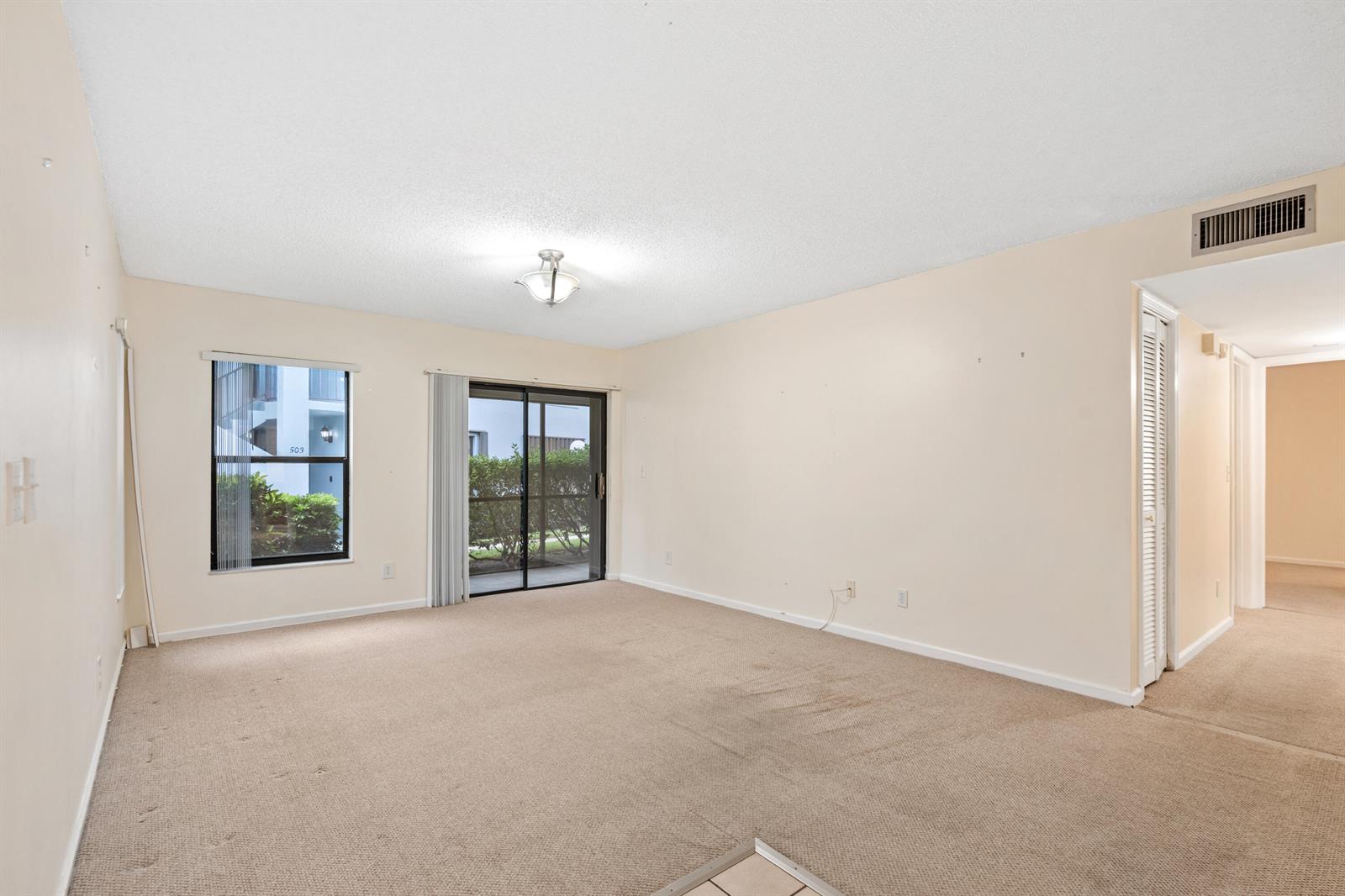 An empty living room features beige carpet, light-colored walls, a sliding glass door, and windows — 950 S Kanner Highway # 402, Stuart FL 34994, STUART, FL