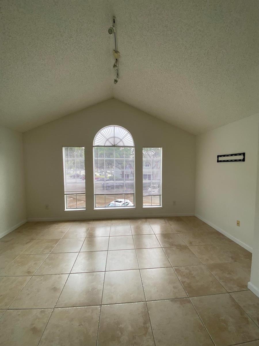 An empty living room features vaulted ceilings, beige tile flooring, and a large arched window — 1020 Crystal Way # H, Delray Beach FL 33444, DELRAY BEACH, FL