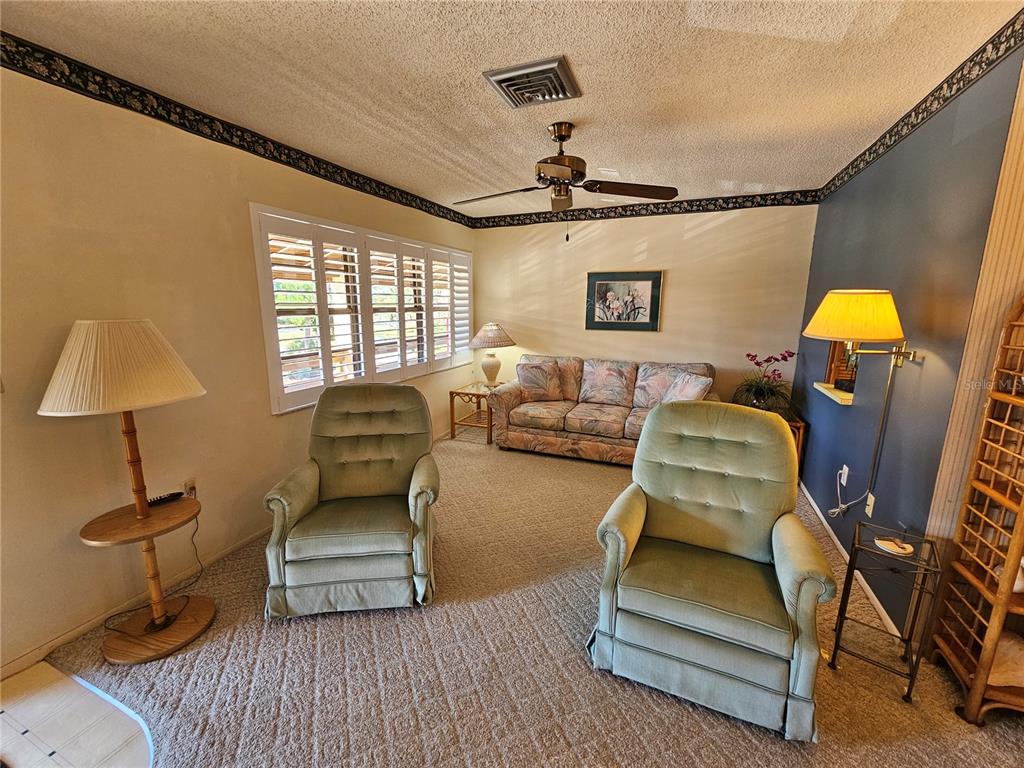 A living room features two green armchairs, a floral sofa, carpeted floors, and plantation shutters — 6 QUAILS RUN BOULEVARD UNIT 9, ENGLEWOOD, FL