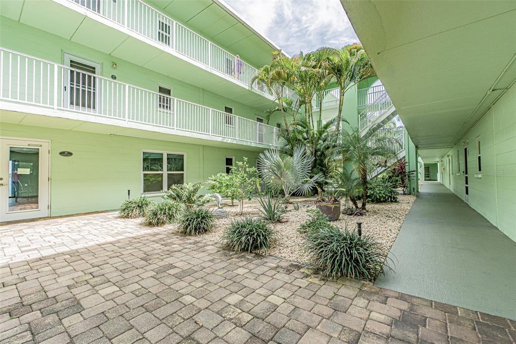 A paved courtyard with tropical landscaping sits between two levels of a green apartment building — 464 PAULA DRIVE N UNIT 116, DUNEDIN, FL