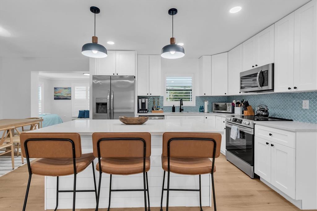 A modern kitchen features white cabinets, a large island with three stools, and blue backsplash — 4986 WOODLAND DRIVE, ST PETERSBURG, FL
