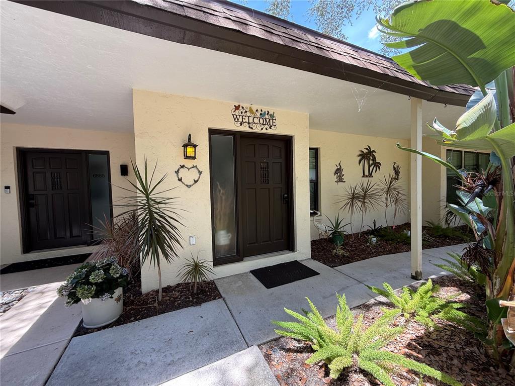 The exterior of a home features a brown front door, a welcome sign, and landscaping — 6861 WOODWIND DR UNIT 75, SARASOTA, FL