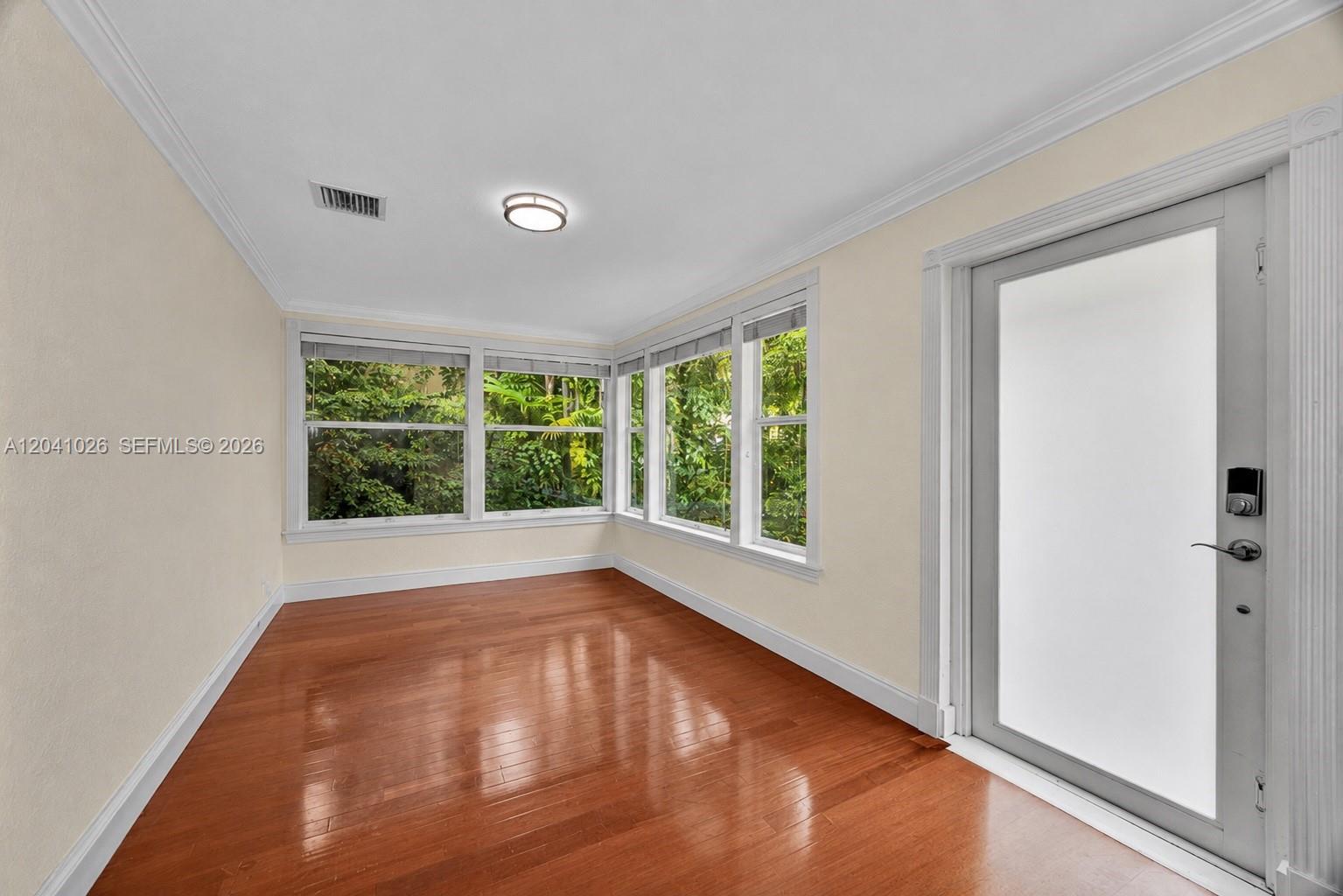 An empty room features hardwood floors, white walls, crown molding, and large windows overlooking greenery — 829 Medina Ave, Coral Gables FL 33134, CORAL GABLES, FL
