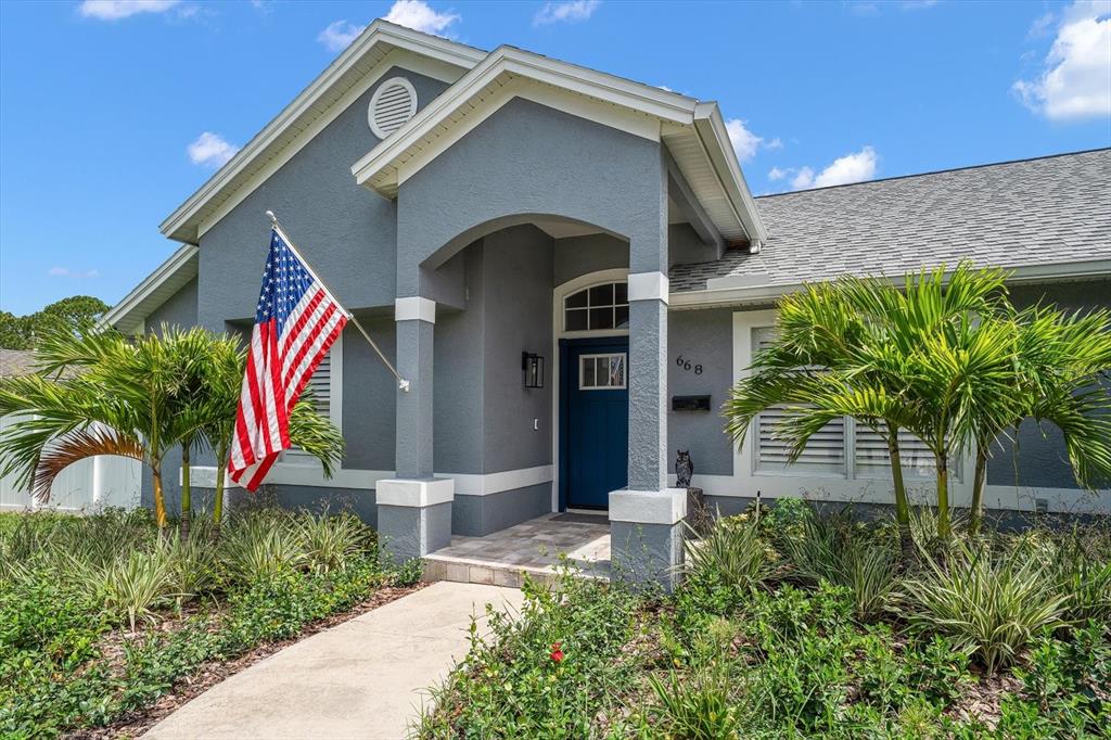 A gray house exterior features a blue front door, a flag, and palm trees — 668 MEHLENBACHER ROAD, BELLEAIR, FL