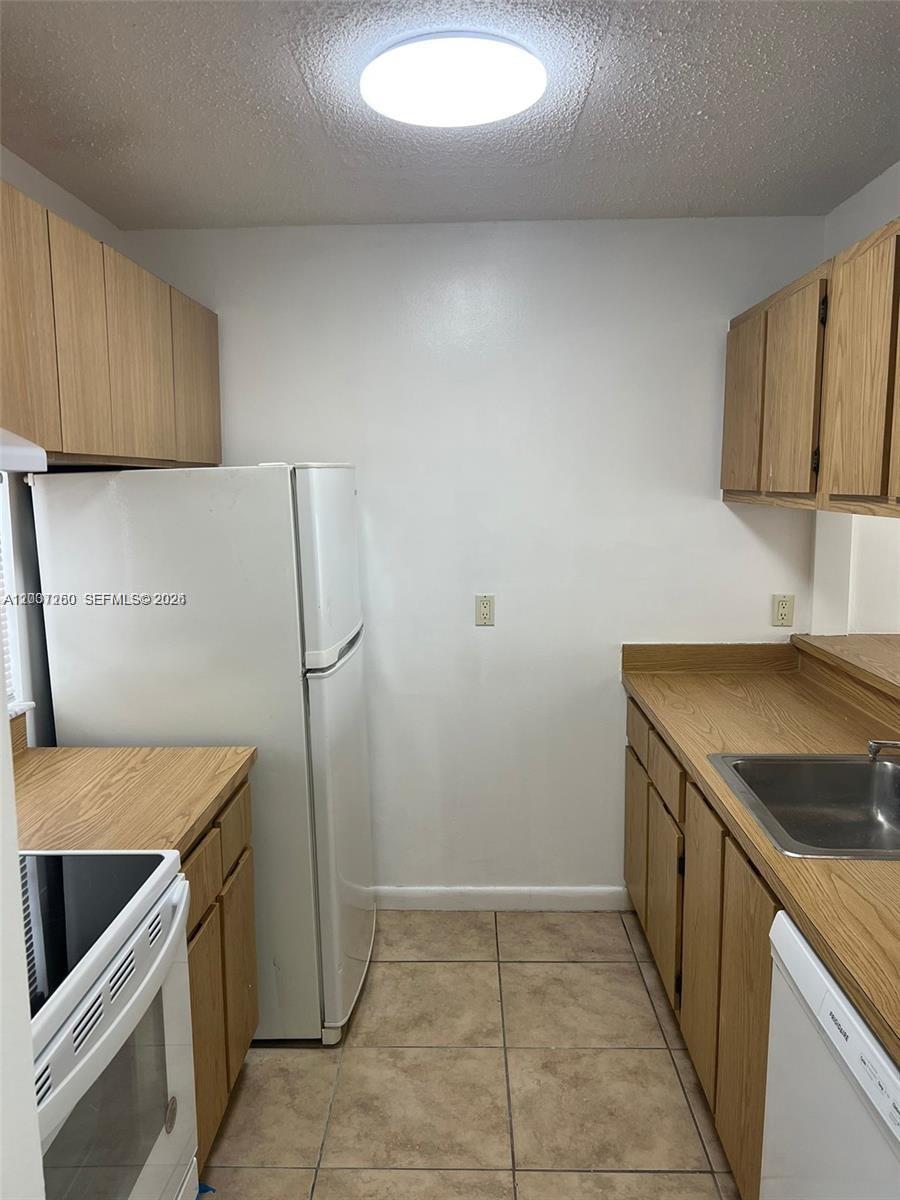 A galley kitchen features light wood cabinets, white appliances, and tan tile flooring — 8610 N Sherman Cir # 504, Miramar FL 33025, MIRAMAR, FL