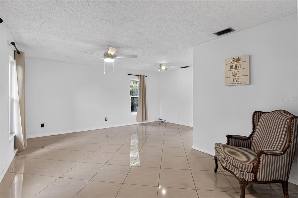 An empty living room features beige tile flooring, white walls, and a striped accent chair — 13635 BERKSHIRE COURT, GRAND ISLAND, FL