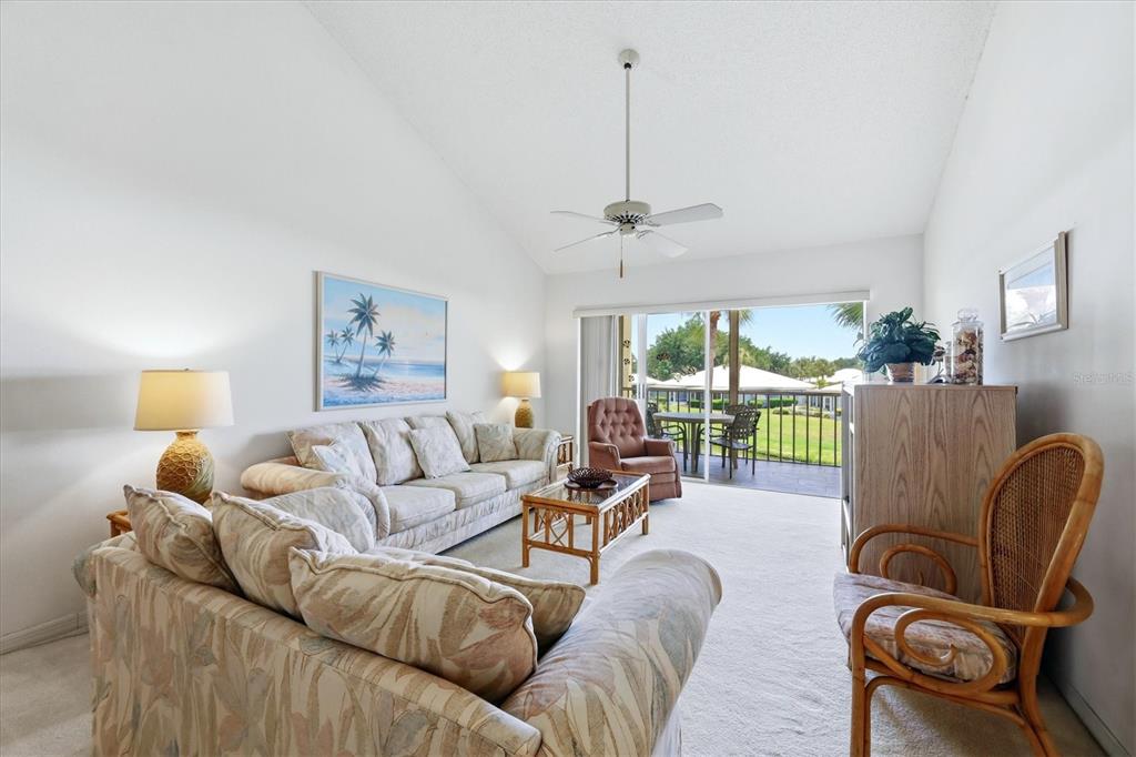 A living room with a vaulted ceiling, beige carpet, tropical-patterned furniture, and a sliding glass door — 444 CERROMAR ROAD UNIT 298, VENICE, FL