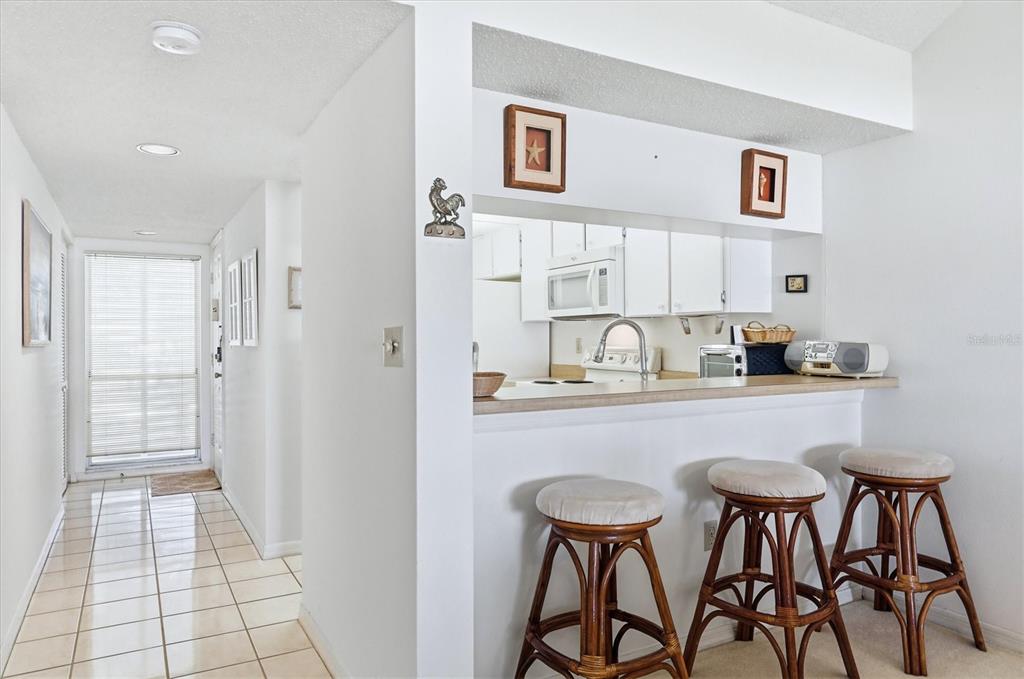 A kitchen with a breakfast bar and stools overlooks a tiled hallway with a door — 444 CERROMAR ROAD UNIT 298, VENICE, FL