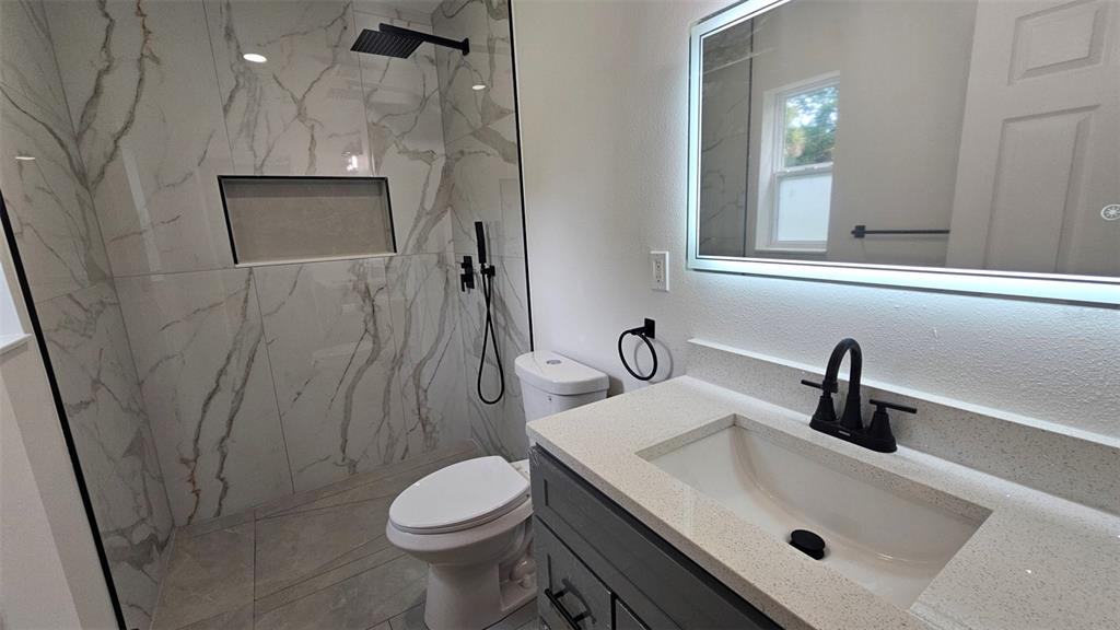 A modern bathroom features a marble-tiled walk-in shower, a gray vanity, and a lighted mirror — 924 FERN AVENUE, COCOA, FL