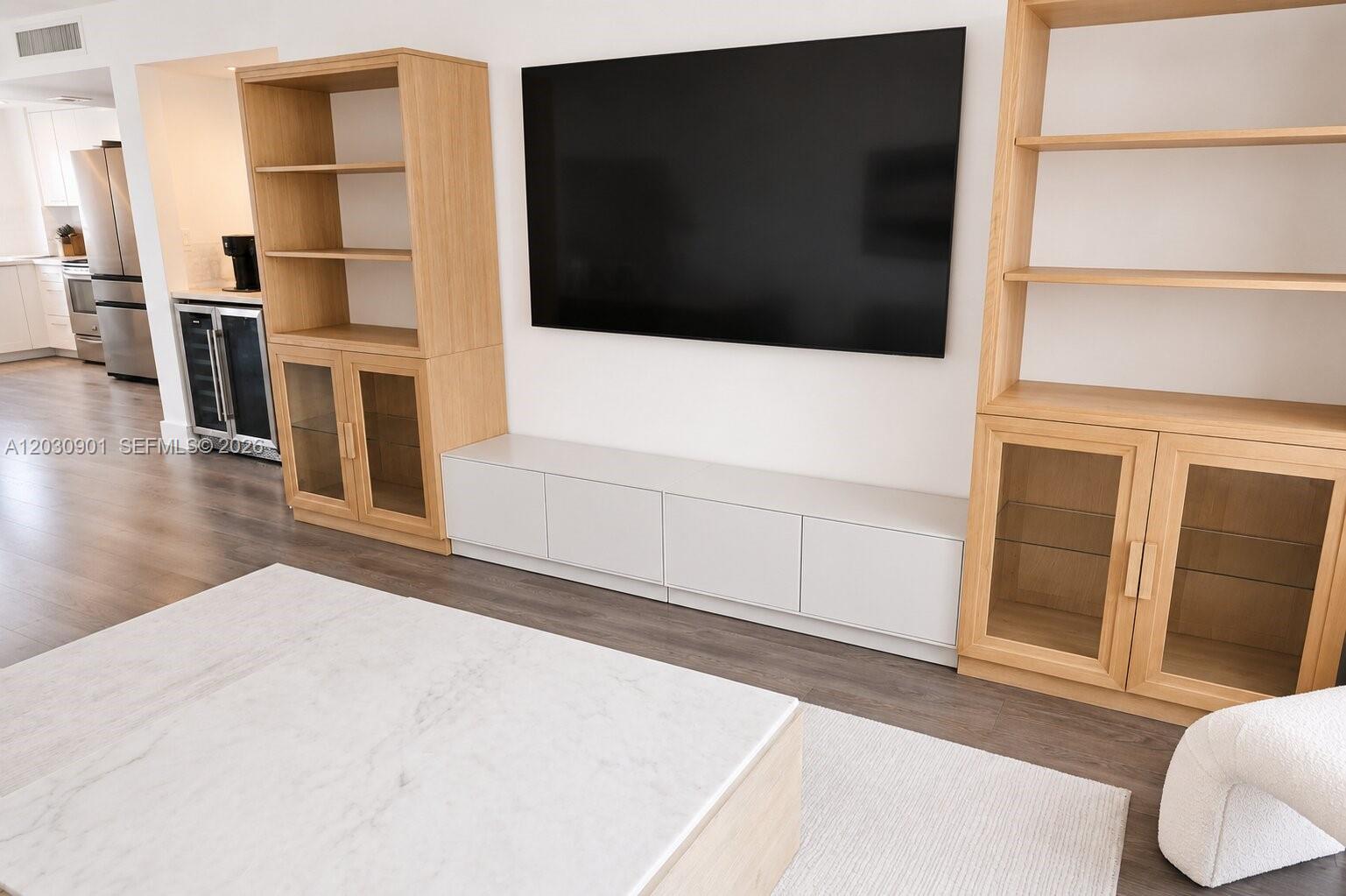 A living room features a wall-mounted television flanked by light wood shelving units — 1627 Brickell Ave # 1005, Miami FL 33129, MIAMI, FL