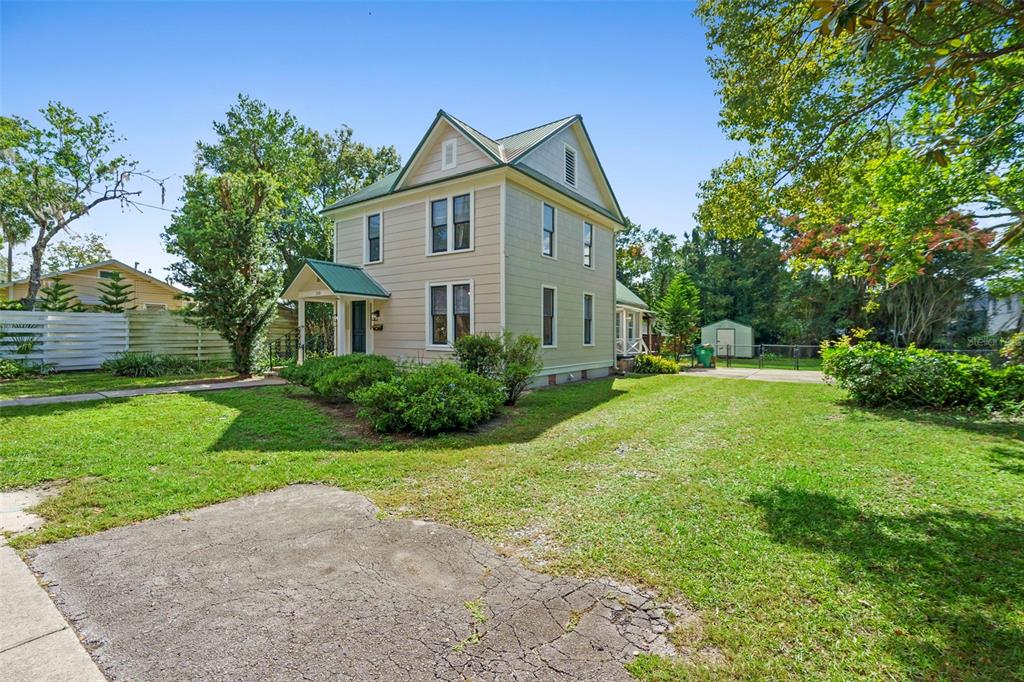 A two-story, light-colored house with a green metal roof stands on a grassy lot — 210 W UNIVERSITY AVENUE, DELAND, FL