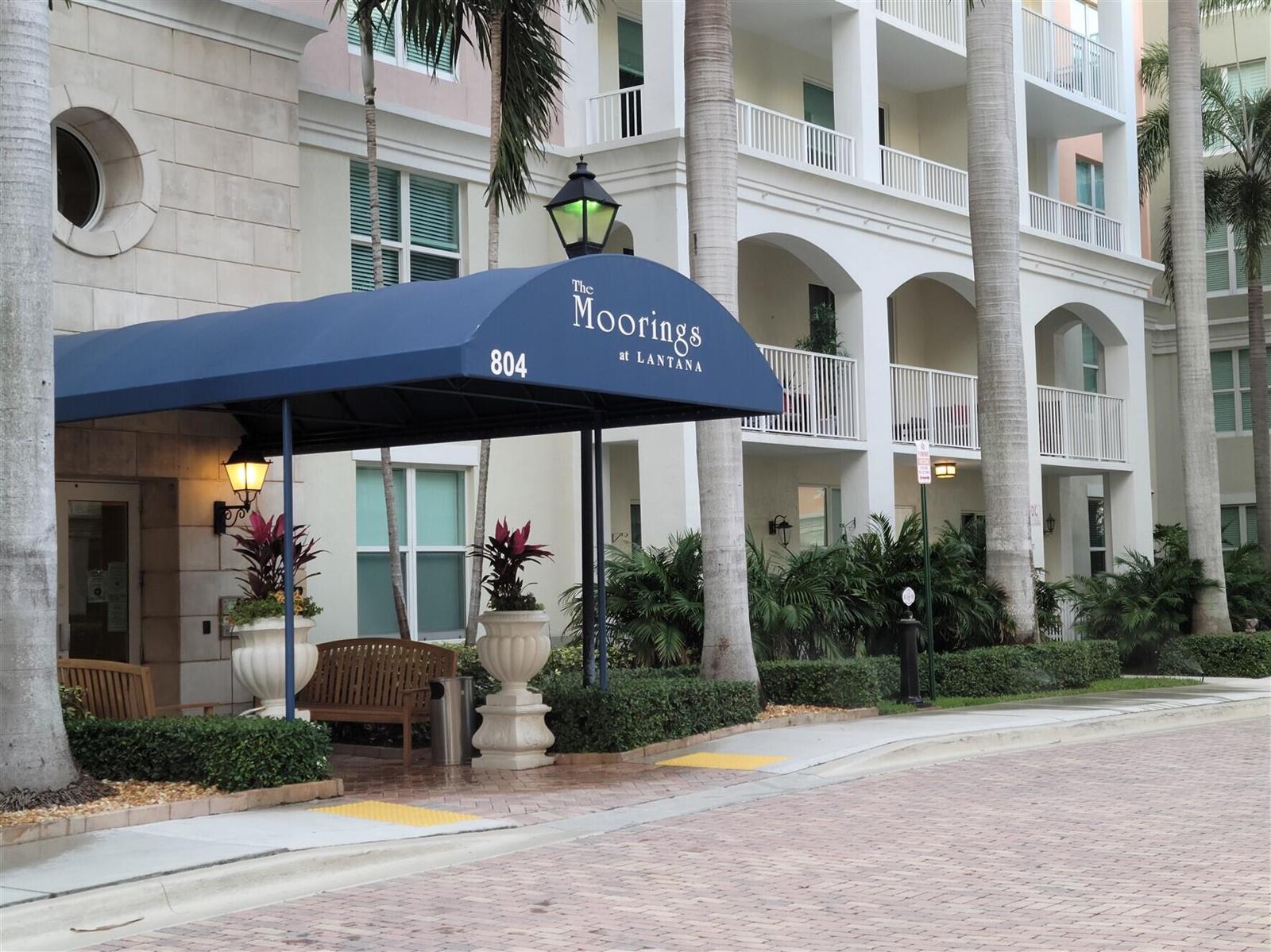 A blue awning covers the entrance to a multi-story building with palm trees and landscaping — 804 E Windward Way # 508, Lantana FL 33462, LANTANA, FL