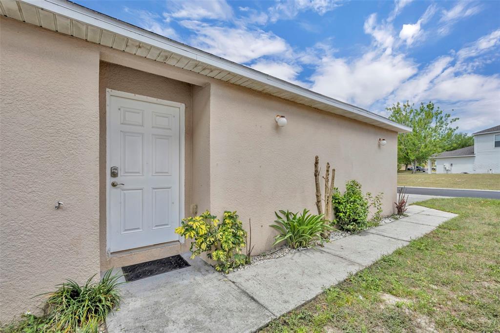 A beige exterior wall features a white door, a concrete walkway, and small landscaping plants — 3147 WHISPERING TRAILS STREET, WINTER HAVEN, FL