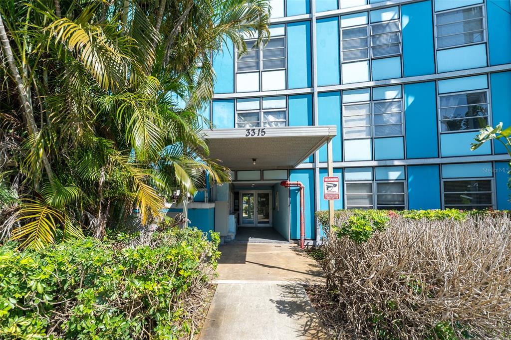 The exterior of a blue multi-story apartment building features a covered entrance and landscaping — 3315 58TH AVENUE S UNIT 512, ST PETERSBURG, FL