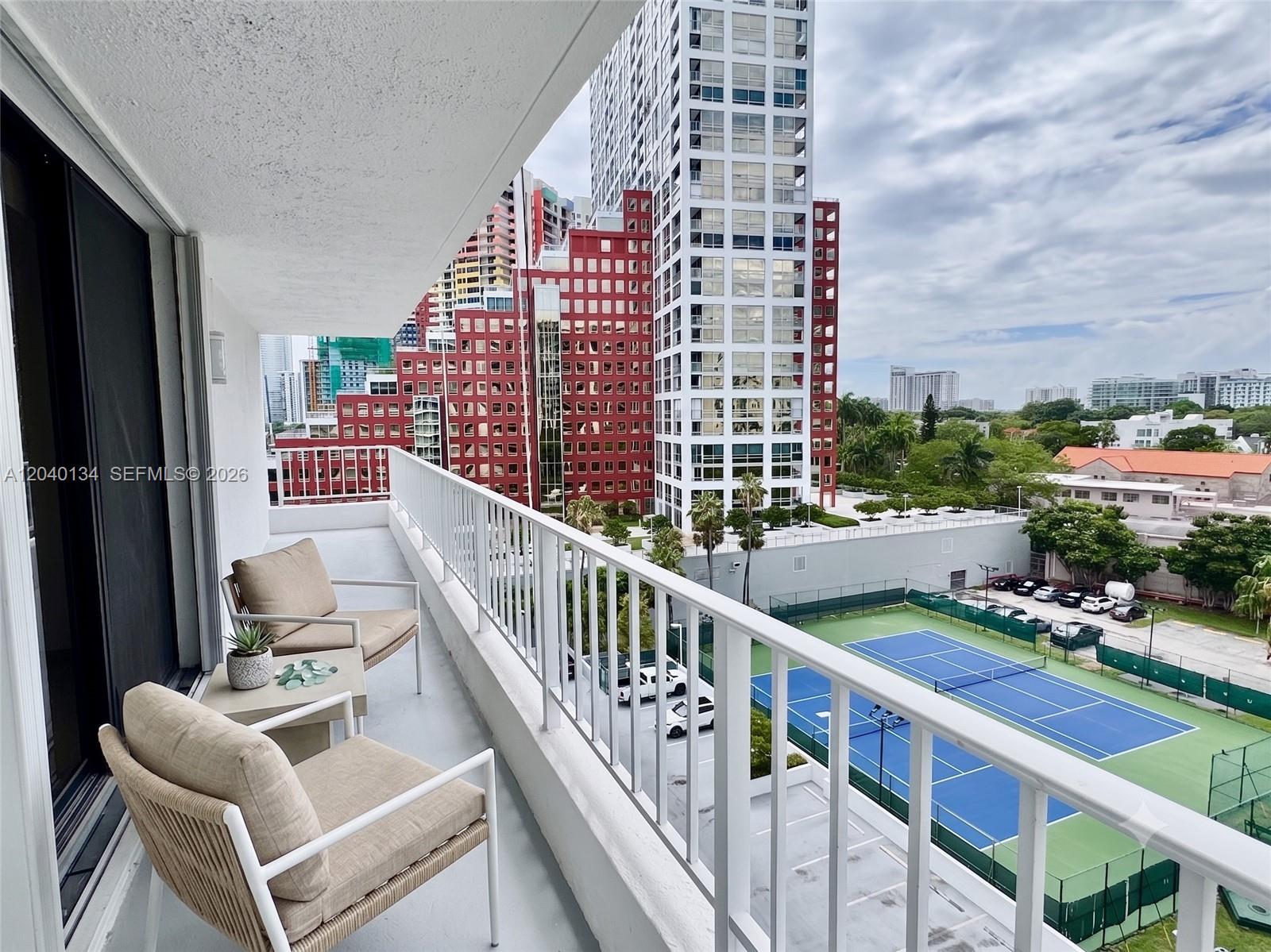 A balcony with two chairs and a small table overlooks a tennis court and city buildings — 200 SE 15th Rd # 9I, Miami FL 33129, MIAMI, FL