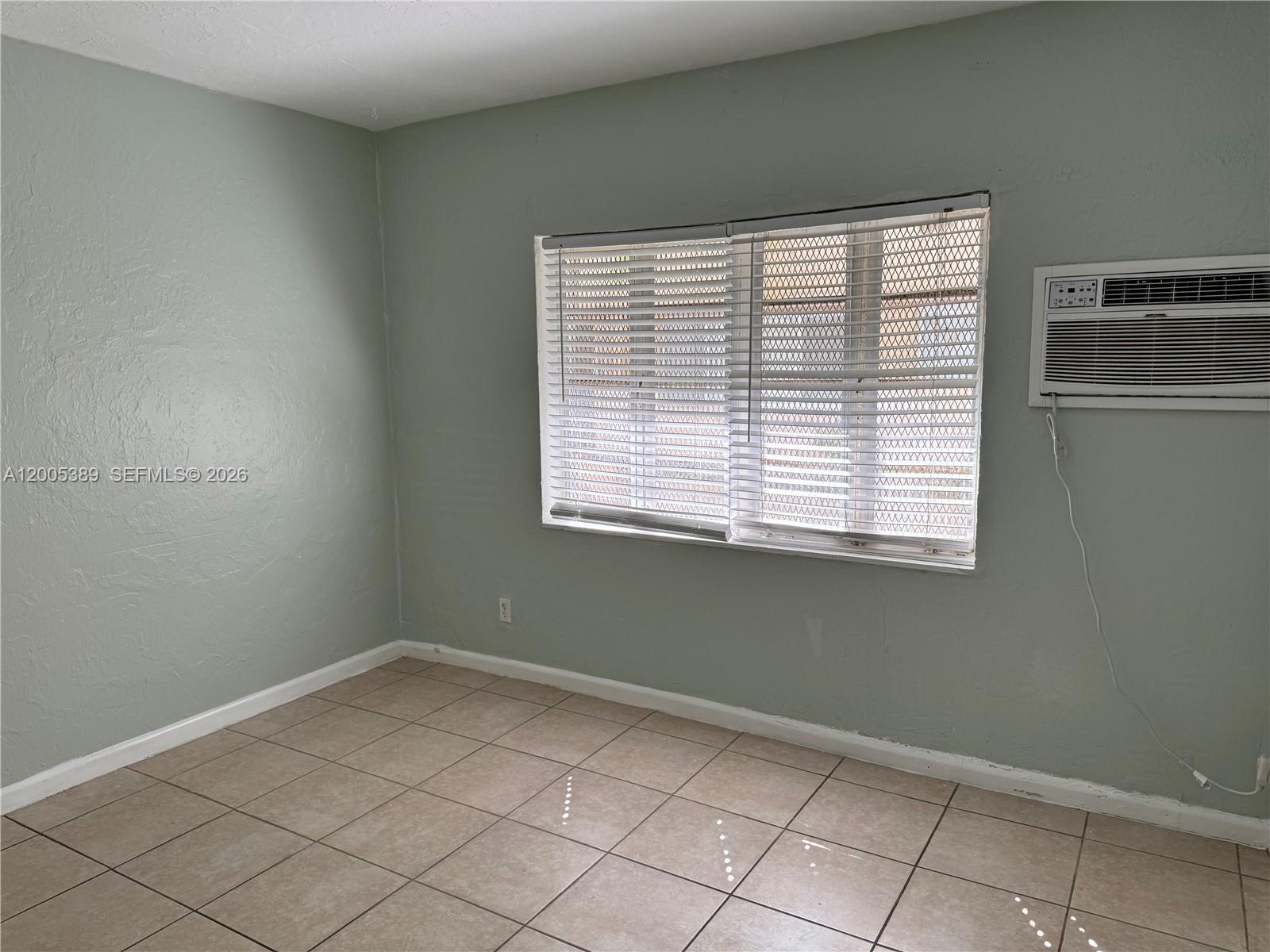 An empty room features light green walls, tiled flooring, a window with blinds, and an air conditioner — 7633 Dickens Ave # 2, Miami Beach FL 33141, MIAMI BEACH, FL