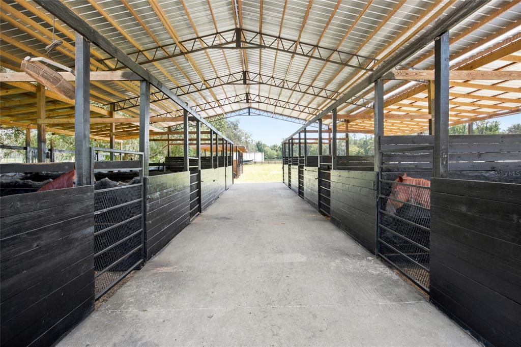 An open-air horse stable features a concrete aisle between rows of black wooden stalls — 3875 SE 118TH LANE, BELLEVIEW, FL