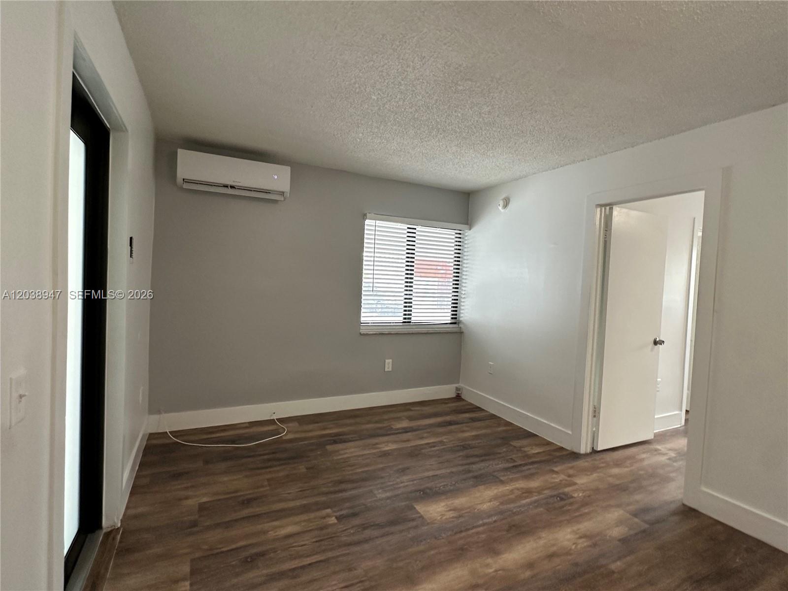 An empty room features wood-look flooring, a wall-mounted air conditioner, a window with blinds, and a doorway — 529 SW 4th St # 104, Miami FL 33130, MIAMI, FL