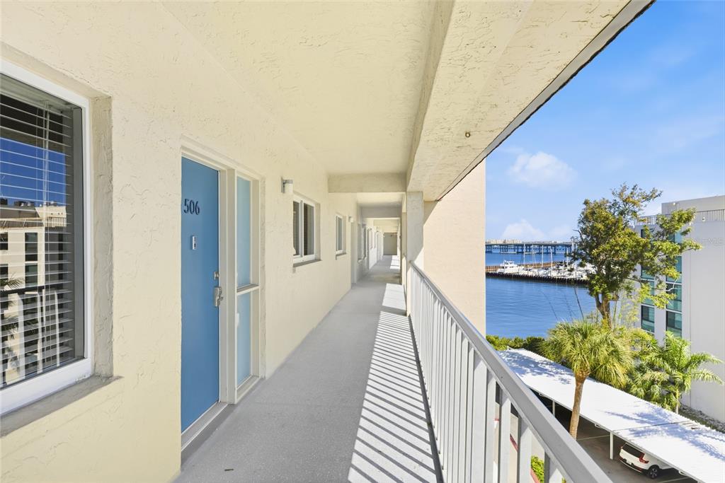 An outdoor walkway with a blue door leads past apartment units toward a waterfront view — 1600 1ST AVENUE W UNIT 506, BRADENTON, FL