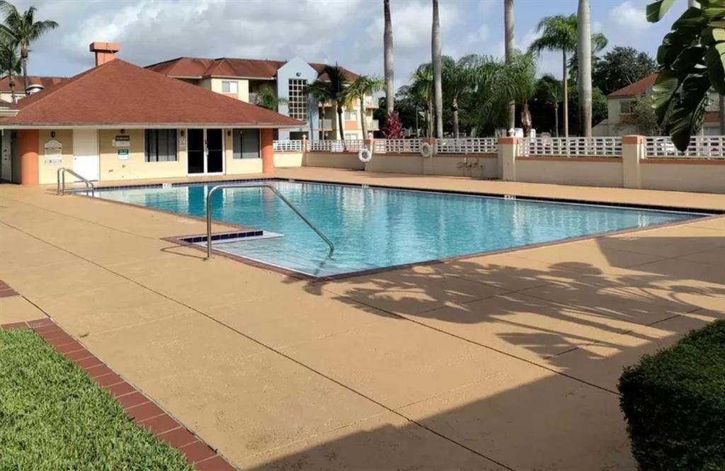 A rectangular swimming pool with a metal handrail sits on a tan patio near buildings — 18860 NW 57th Avenue # 106, Hialeah FL 33015, HIALEAH, FL