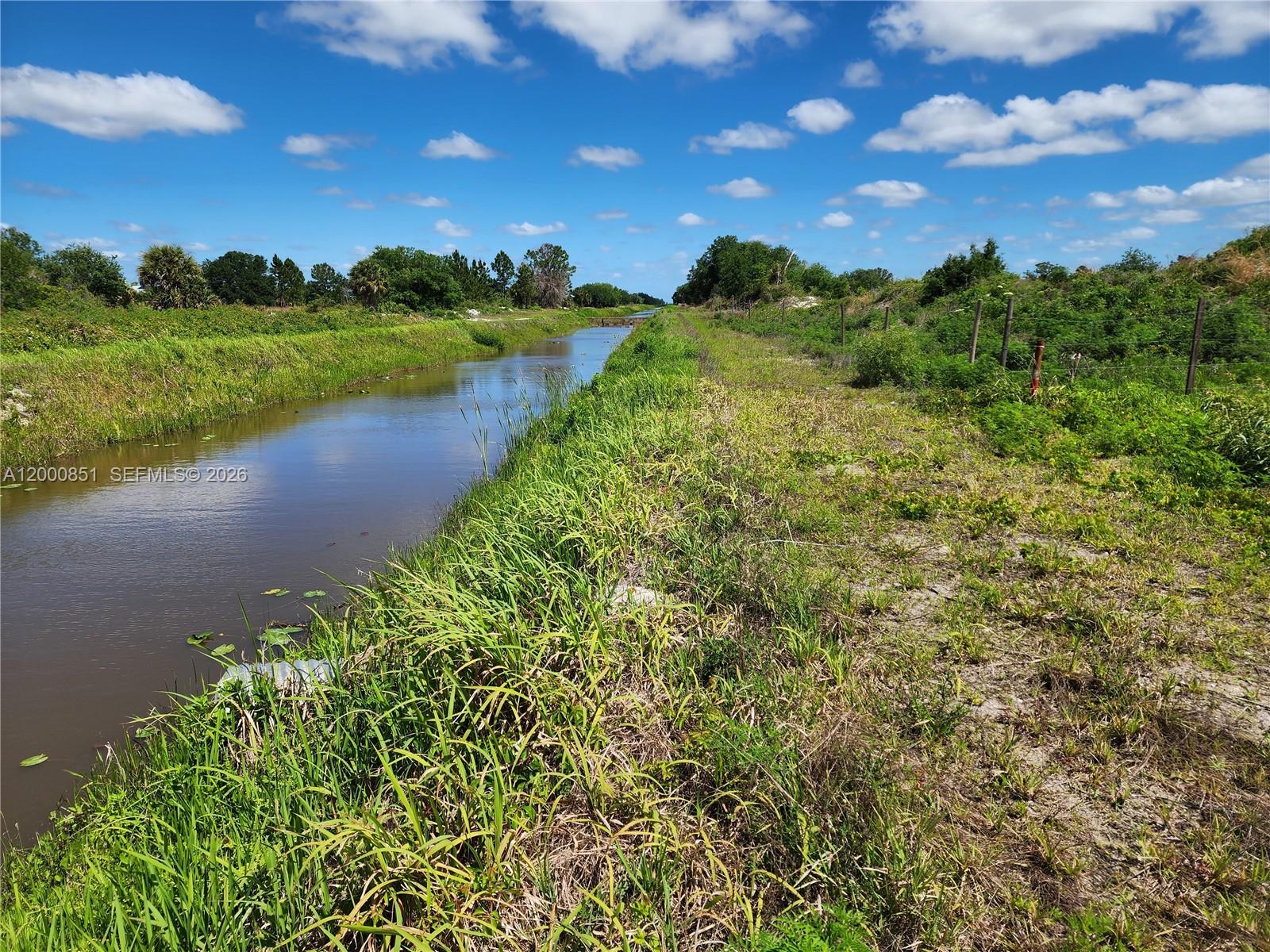 A narrow canal flows through a grassy field under a bright blue sky — 16785 NW 292nd ST, Okeechobee FL 34972, OKEECHOBEE, FL