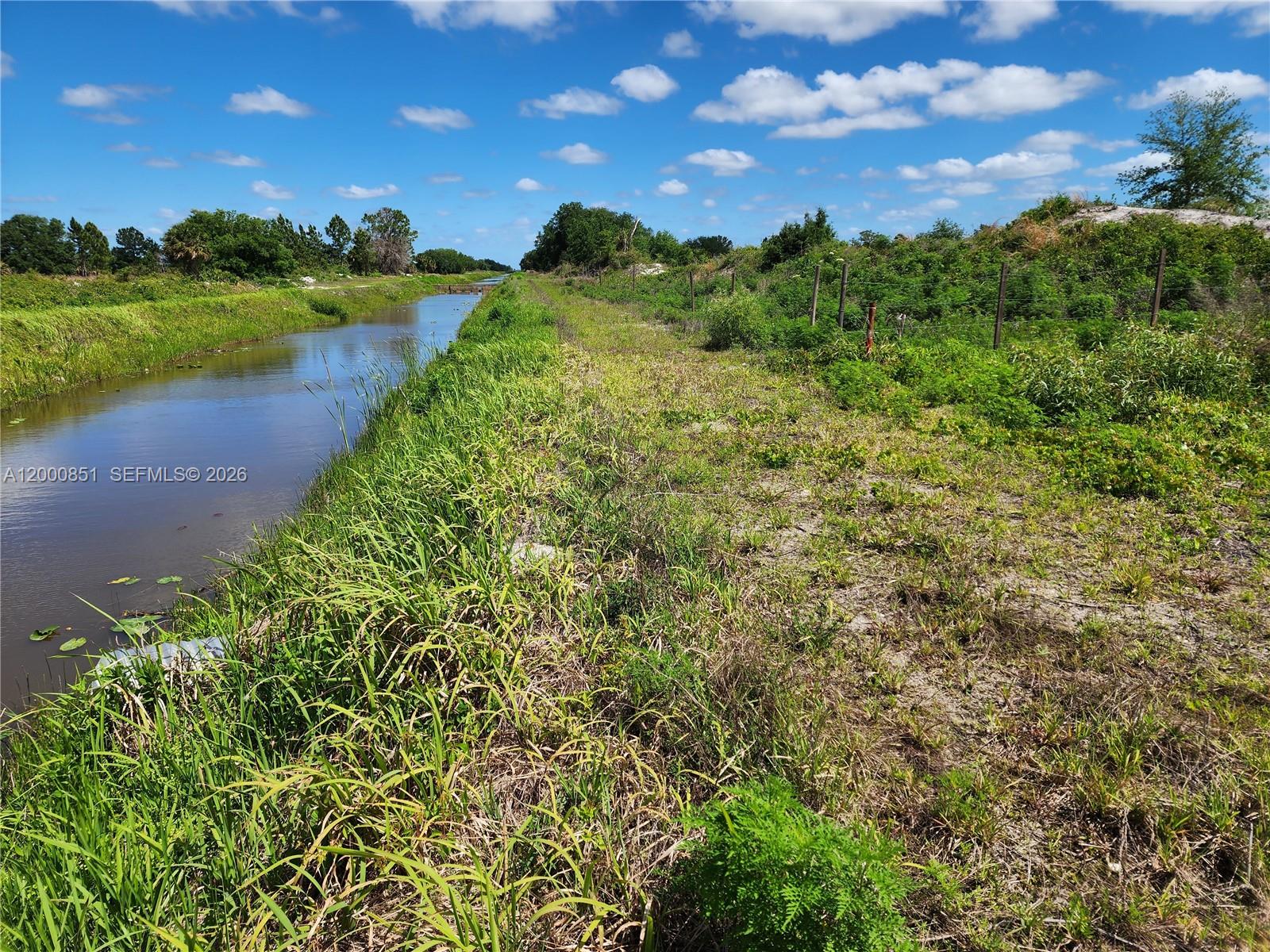 A narrow canal runs alongside a grassy bank with a wire fence under a blue sky — 16785 NW 292nd ST, Okeechobee FL 34972, OKEECHOBEE, FL