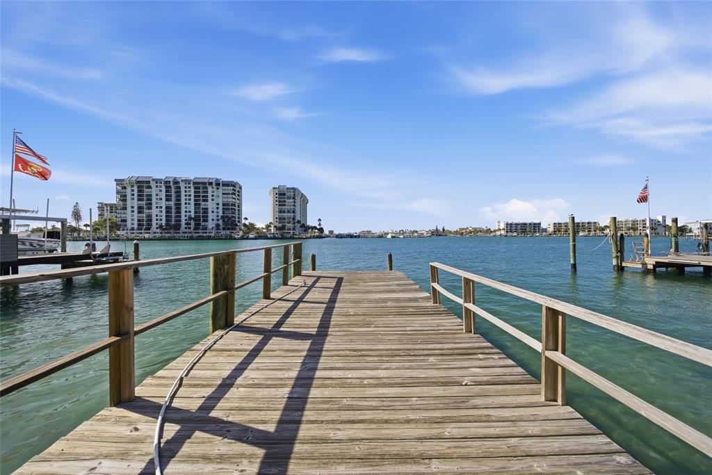 A wooden dock extends over calm water toward a distant shoreline with apartment buildings — 4 BELLEVUE DRIVE, TREASURE ISLAND, FL