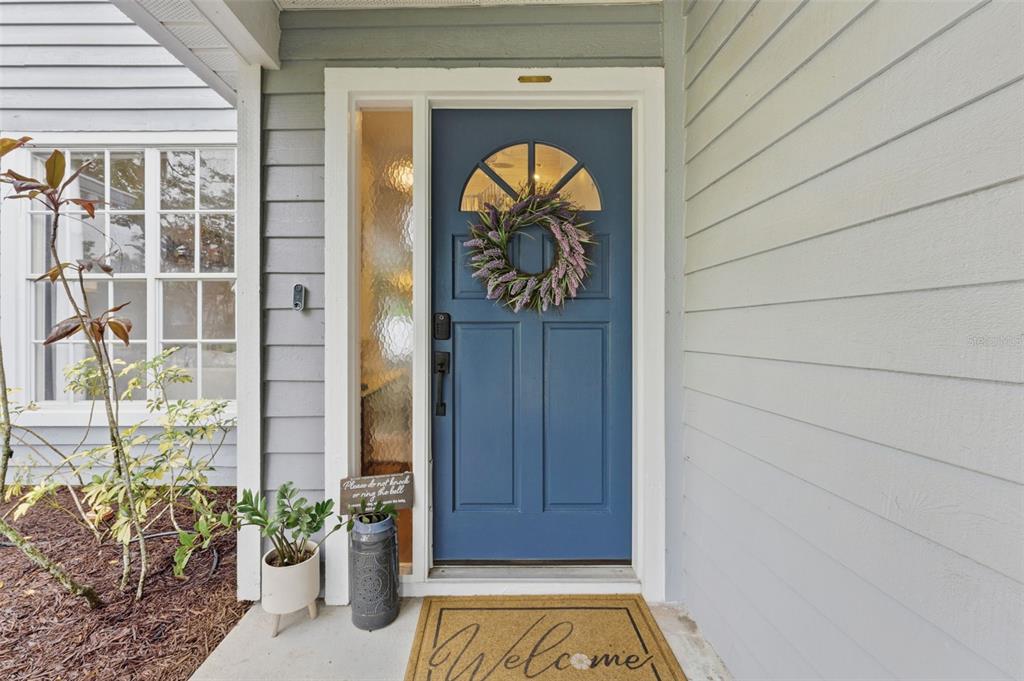 A blue front door with a wreath and a welcome mat sits on a covered porch — 7226 HUMMINGBIRD LANE, NEW PORT RICHEY, FL
