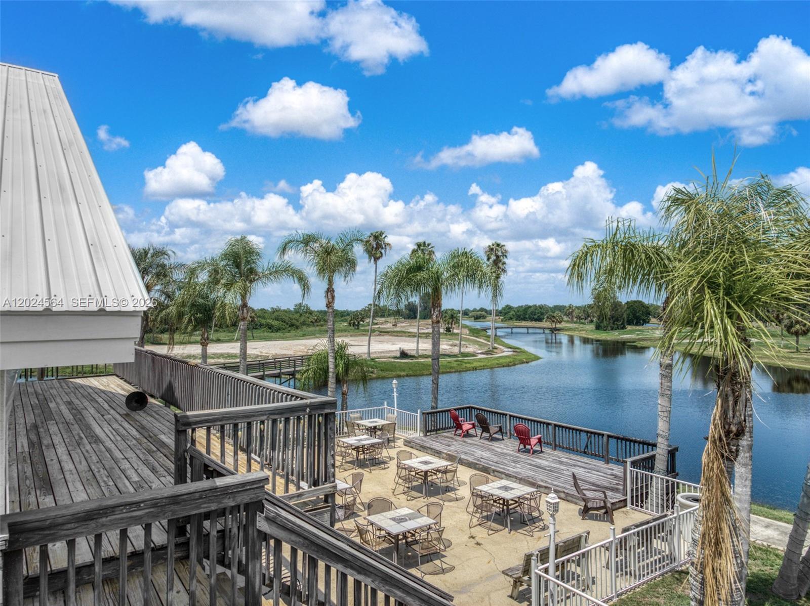 An elevated wooden deck overlooks a patio with tables and a lake with palm trees — 6813 Heavitree, Sebring FL 33876, SEBRING, FL