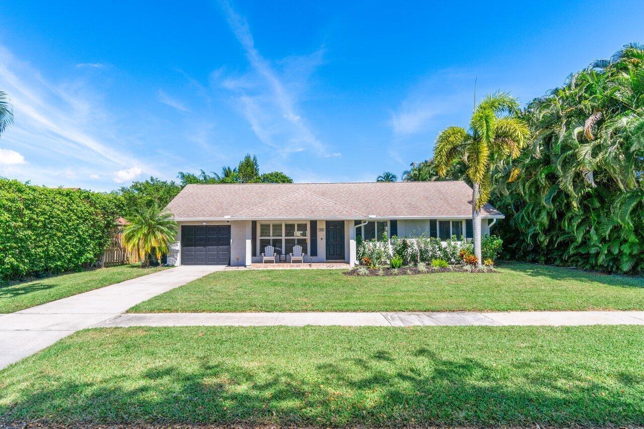 A single-story house with a gray garage, front lawn, and lush landscaping under a blue sky — 1315 Fulmar Drive, Delray Beach FL 33444, DELRAY BEACH, FL