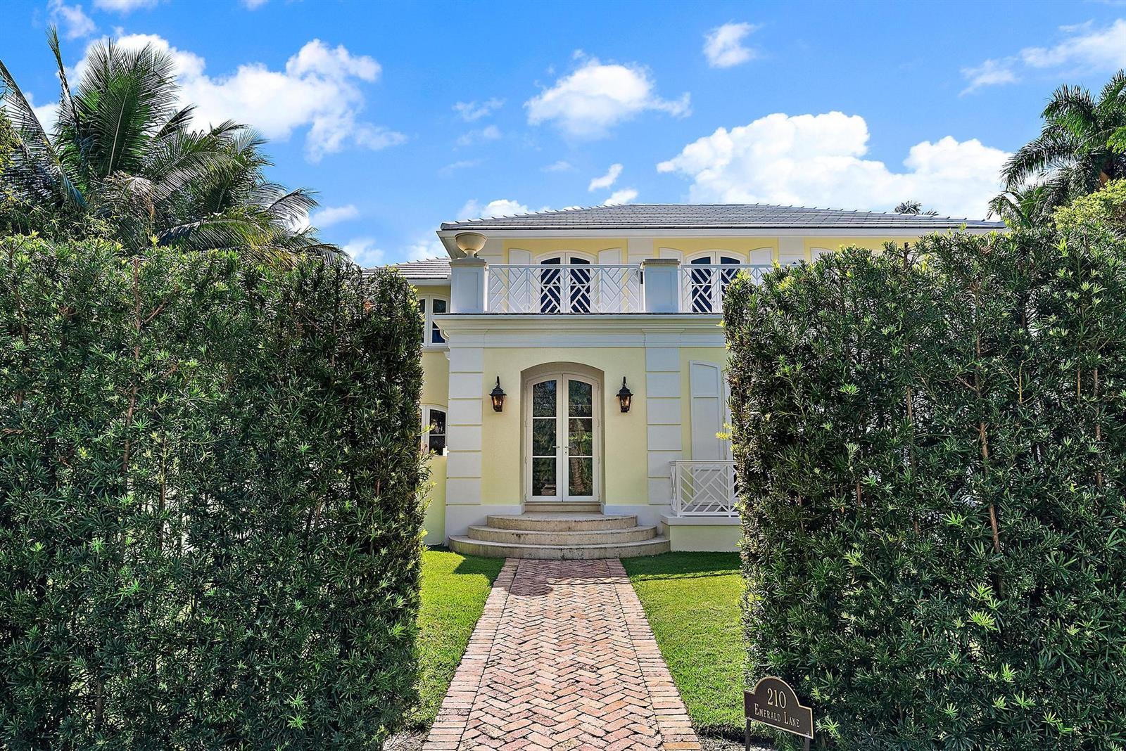 A yellow two-story house with a brick walkway is framed by tall green hedges — 210 Emerald Lane, Palm Beach FL 33480, PALM BEACH, FL