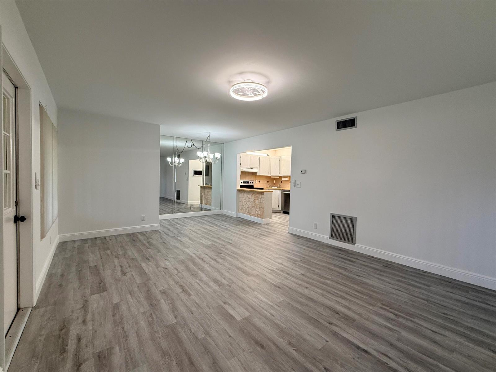 An empty living room with gray wood-look flooring, a mirrored wall, and an open kitchen — 318 Knotty Pine Circle # 2, FL 33463, FL