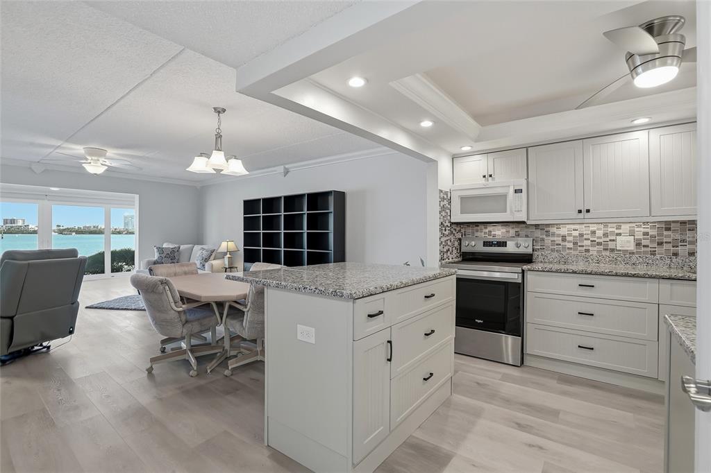 A kitchen with light gray cabinetry and granite countertops opens to a dining and living area — 868 BAYWAY BOULEVARD UNIT 101, CLEARWATER BEACH, FL