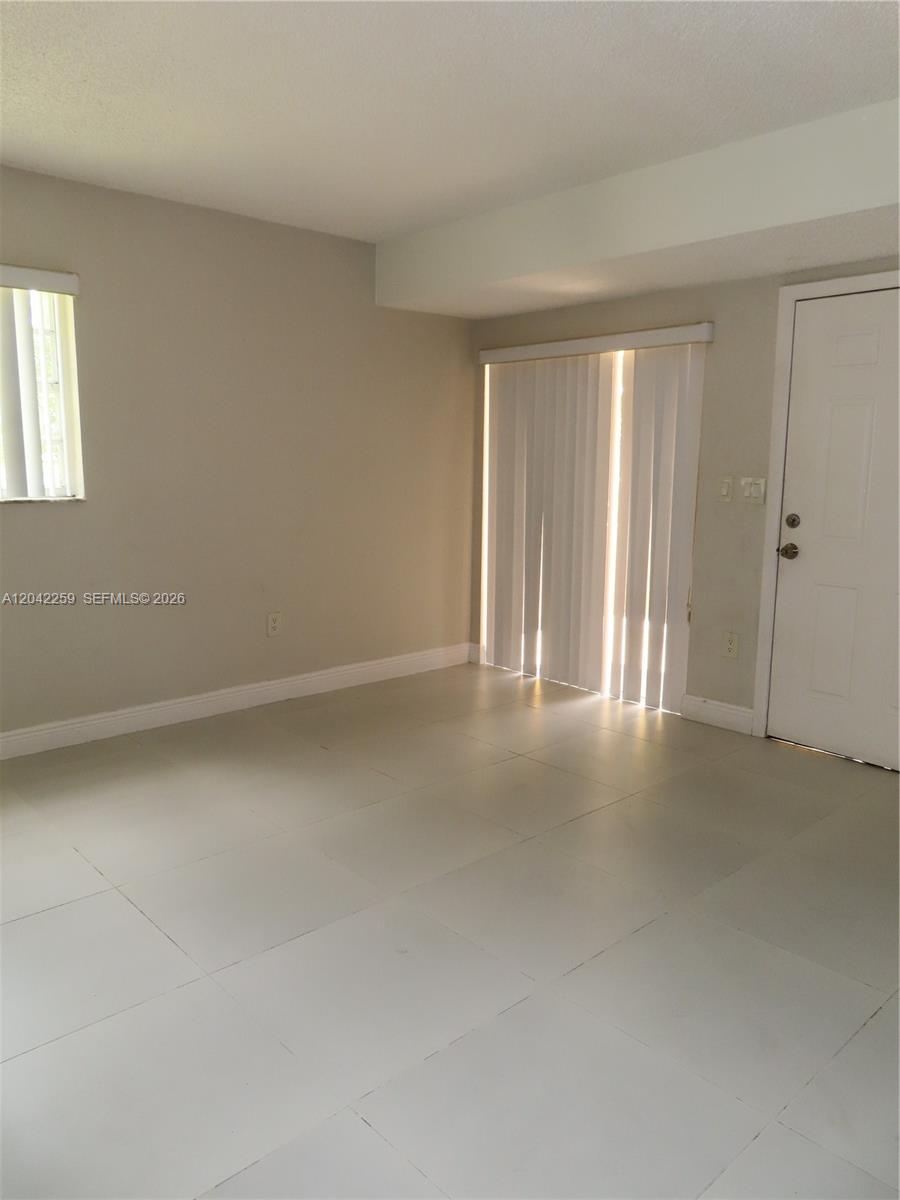 An empty room features light-colored tile flooring, beige walls, a window, and sliding glass doors — 13894 SW 65th Ter # 12, Miami FL 33183, MIAMI, FL