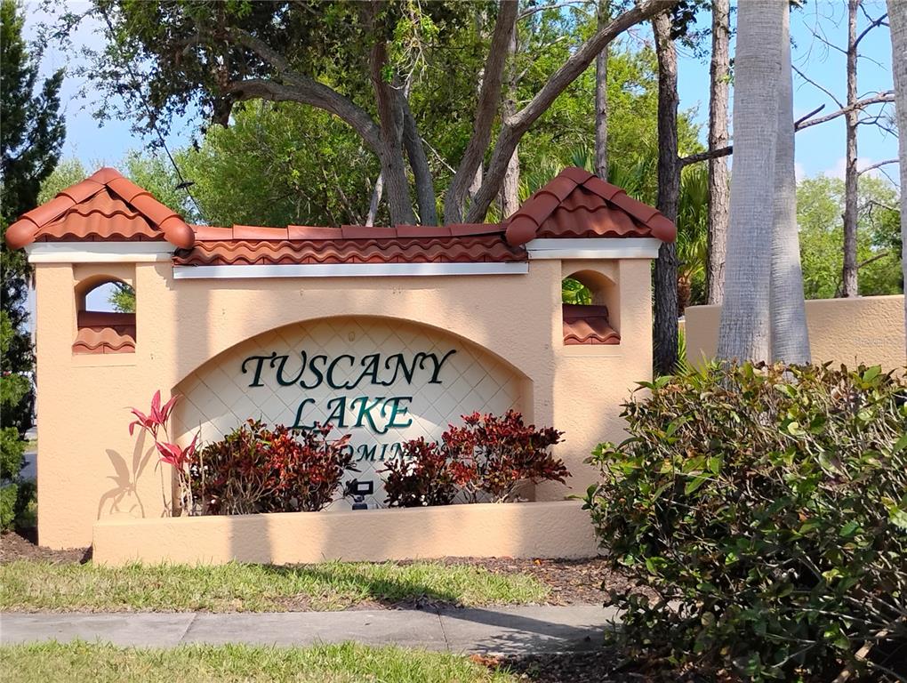 A stucco monument sign for Tuscany Lake stands outdoors surrounded by trees and landscaping — 1761 AUBURN LAKES DRIVE UNIT 11, VENICE, FL