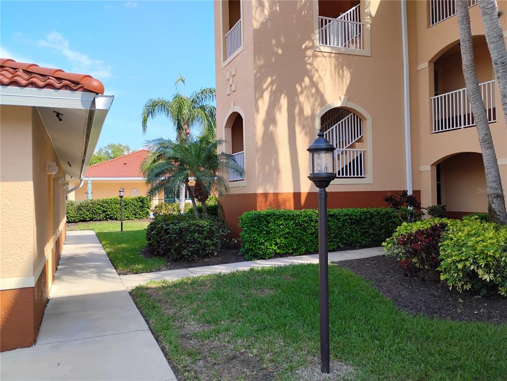 A paved walkway leads past a tan apartment building with balconies and landscaped bushes — 1761 AUBURN LAKES DRIVE UNIT 11, VENICE, FL