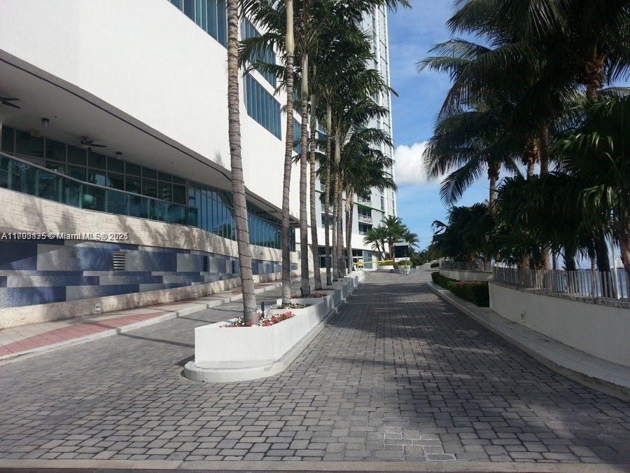 A paved driveway leads toward a modern building lined with palm trees and white planters — 325 S Biscayne Blvd # 3822, Miami FL 33131, MIAMI, FL
