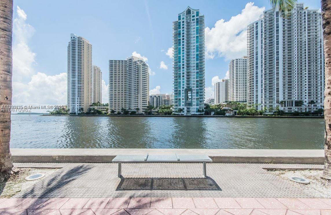 A metal bench sits on a paved waterfront walkway facing a skyline of tall buildings — 325 S Biscayne Blvd # 3822, Miami FL 33131, MIAMI, FL
