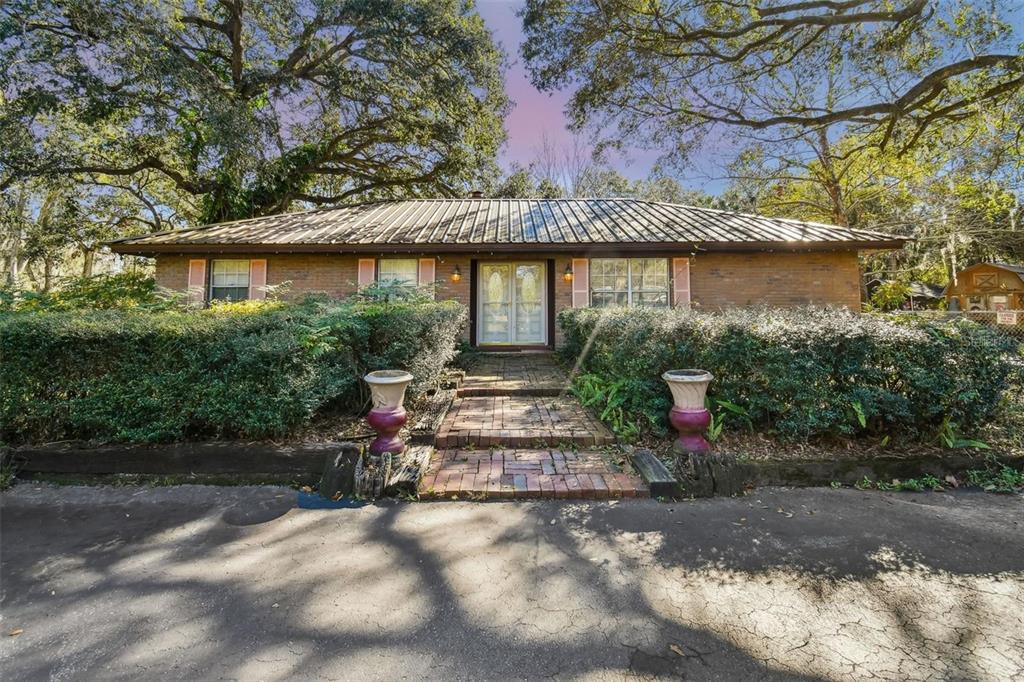 A single-story brick house features a metal roof, a brick walkway, and large shade trees — 10901 MCMULLEN LOOP, RIVERVIEW, FL