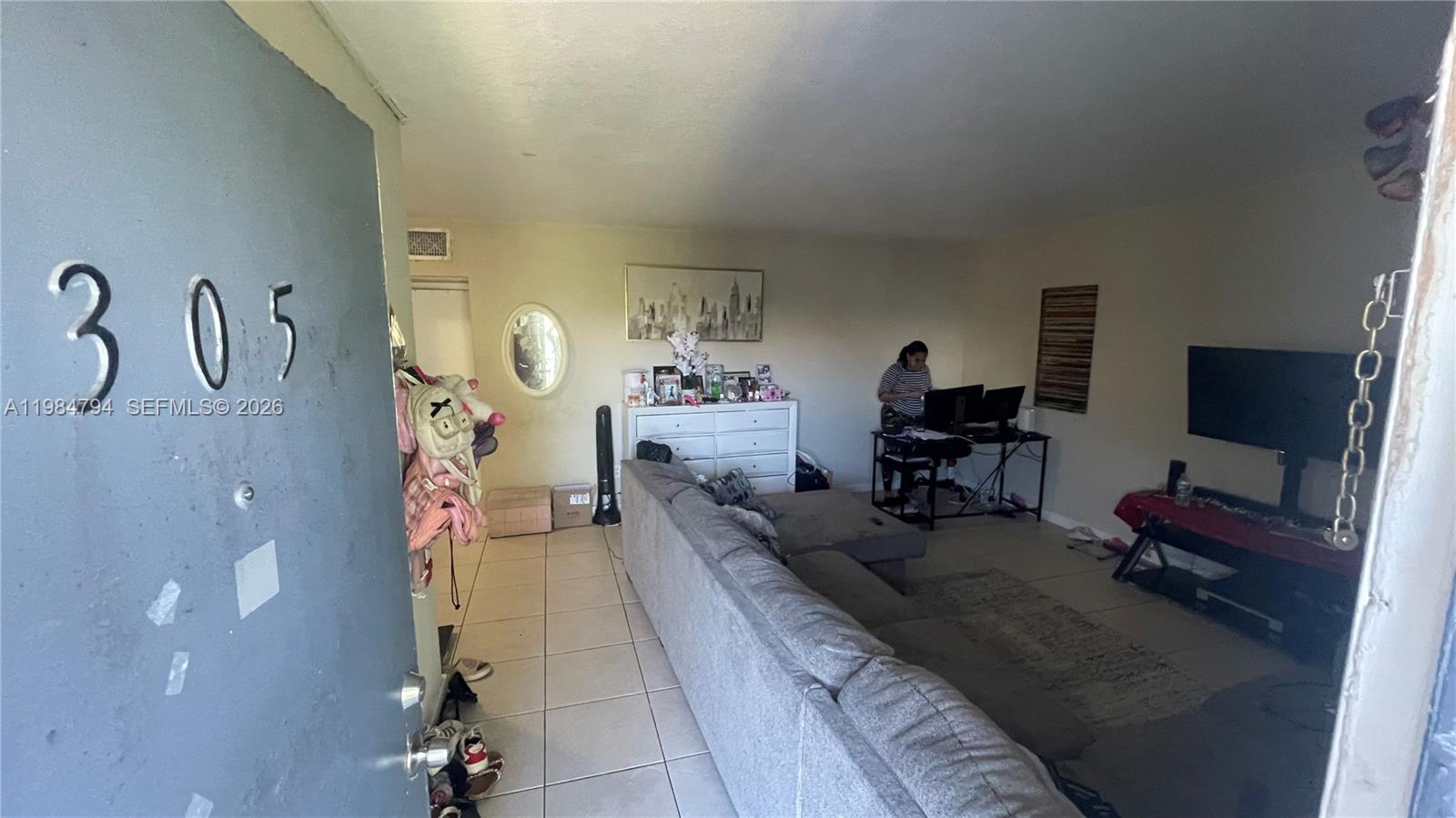 A living room features a gray sectional sofa, a desk with a computer, and tile flooring — 13700 NE 6th Ave # 305, North Miami FL 33161, NORTH MIAMI, FL