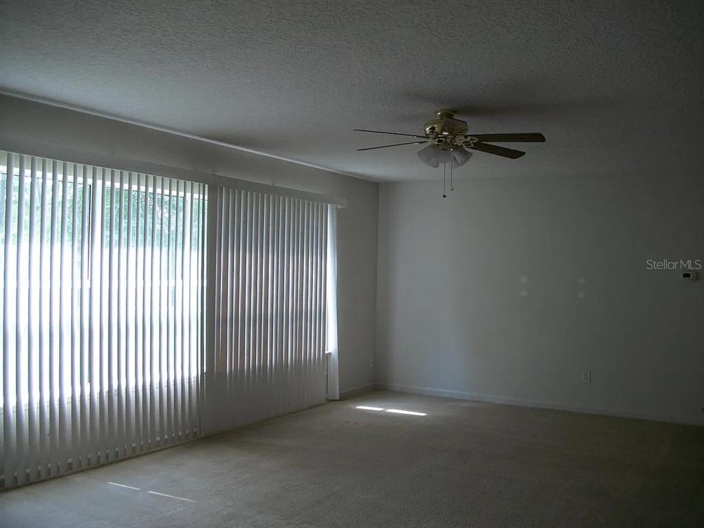 A carpeted living room features a ceiling fan and large windows with vertical blinds — 72 ROLLINS LANE, PALM COAST, FL