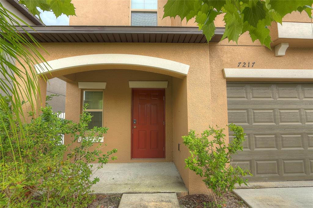 The exterior of a two-story tan stucco home features a red front door and garage — 7217 RONNIE GARDENS COURT, TAMPA, FL