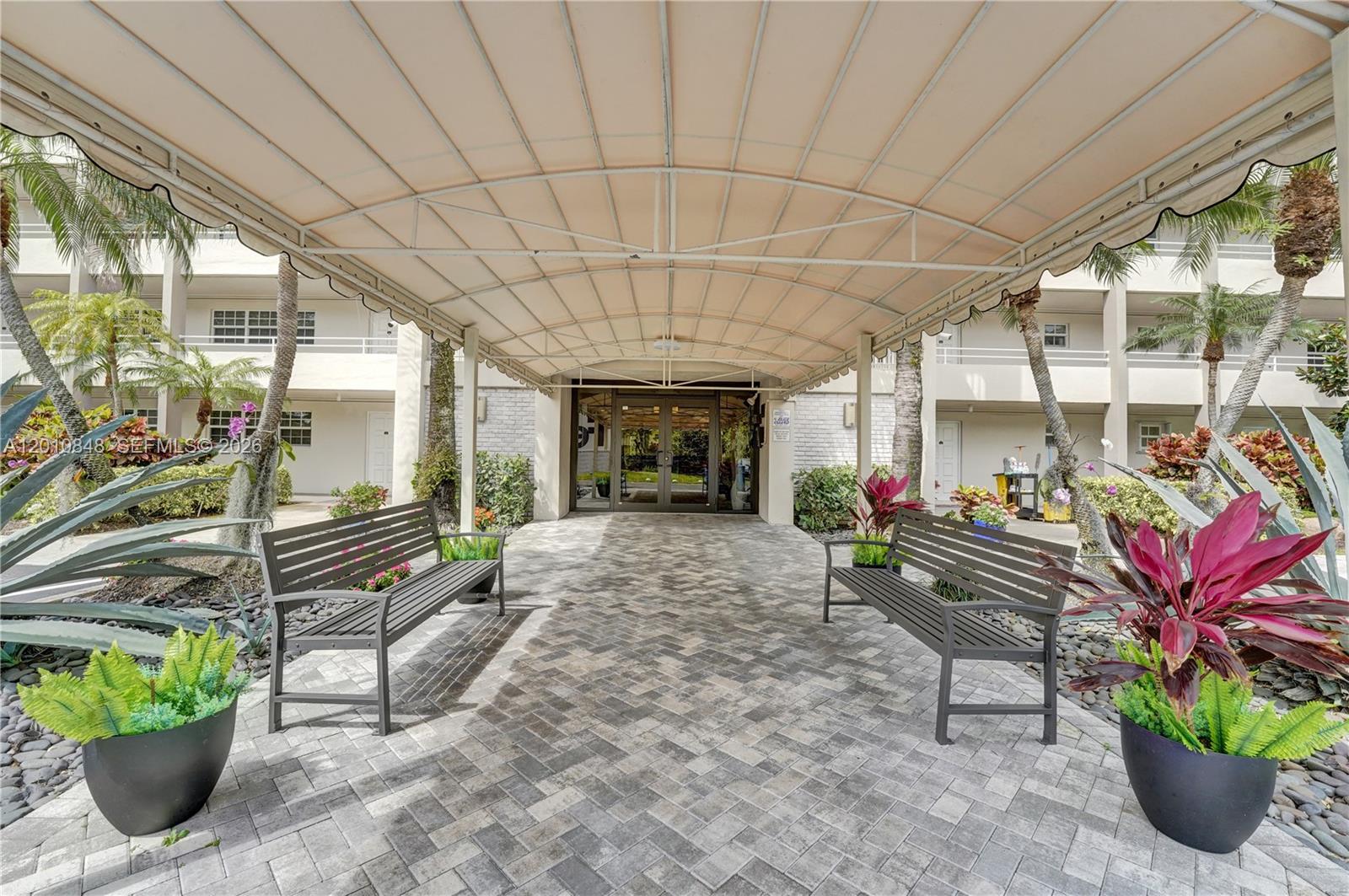 A covered walkway leads to a building entrance flanked by two benches and potted plants — 575 Oaks Ln # 508, Pompano Beach FL 33069, POMPANO BEACH, FL