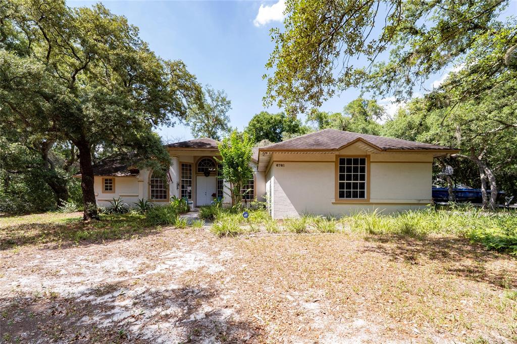 A single-story house with a light-colored exterior sits among mature trees and sandy landscaping — 6781 W LIVINGSTON STREET, ORLANDO, FL