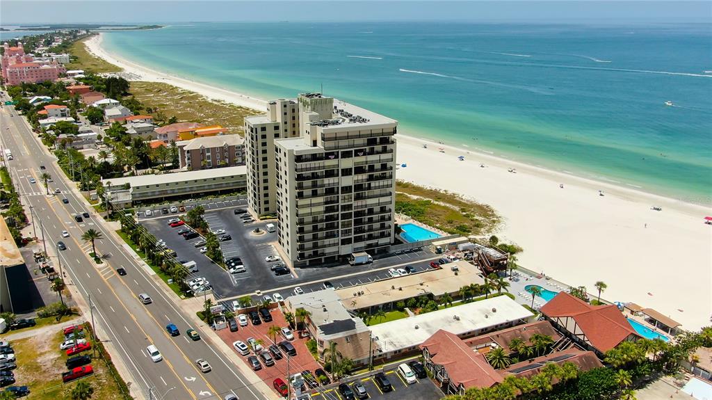 An aerial view shows a high-rise building, parking lots, and swimming pools near a beach — 3820 GULF BOULEVARD UNIT 608, ST PETE BEACH, FL
