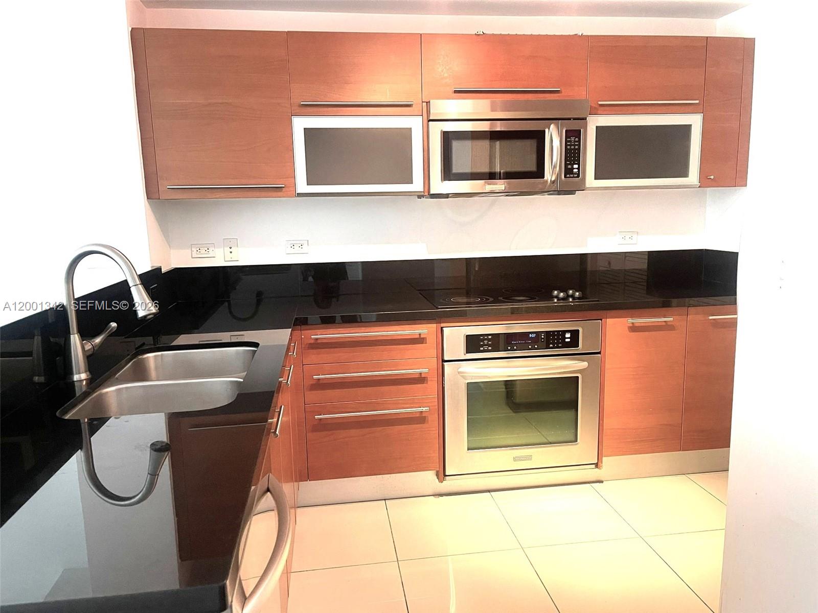 A kitchen features wood-toned cabinets, stainless steel appliances, and black countertops over white tile floors — 253 NE 2nd St # 1004, Miami FL 33132, MIAMI, FL