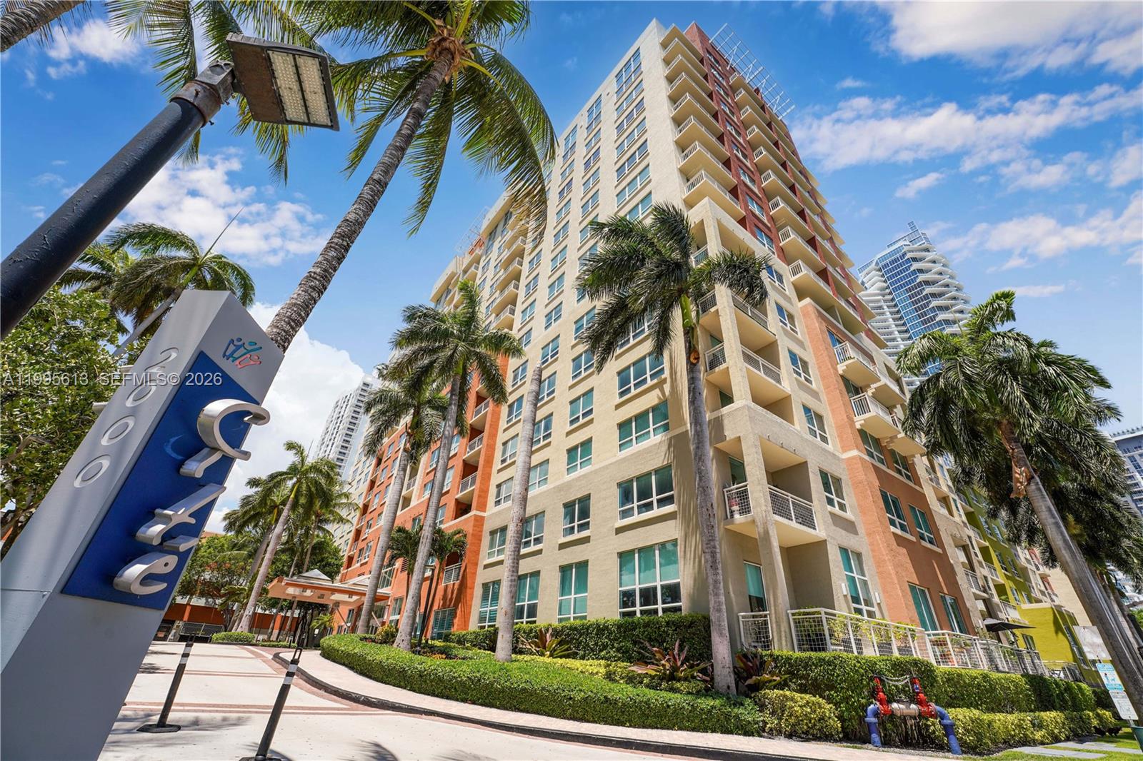 A tall multi-story apartment building with balconies stands behind a sign and palm trees — 2000 N Bayshore Dr # 702, Miami FL 33137, MIAMI, FL